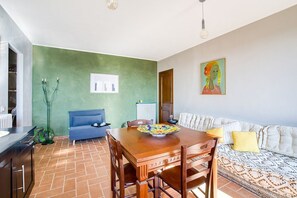 A dining area with a wooden table, chairs, and a bowl. There is a sofa with patterned cushions and a painting on the wall.