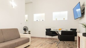 A modern living room with a sofa, dining table, chairs, a TV, and a fan. - GuestHost - Borgata Santa Lucia Cozy Apartment! (Siracusa)