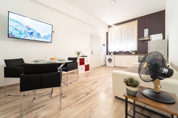 A modern living room with a dining area, a flat-screen TV, a fan, and a kitchen in the background.
