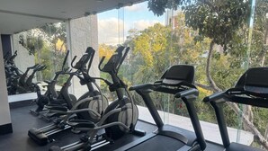 Un gimnasio con varias cintas de correr y bicicletas estáticas, amplias ventanas con vistas a árboles y edificios.