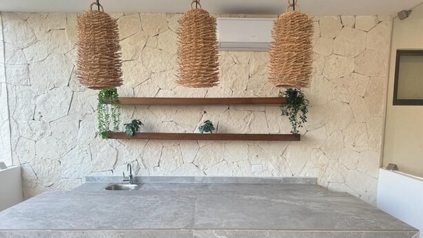 Una cocina con una isla grande, un protector contra salpicaduras de piedra, lámparas de mimbre colgantes y taburetes de madera.