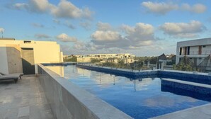 Una piscina en la azotea con agua cristalina y azul, rodeada por una terraza de hormigón y vistas a edificios al fondo.