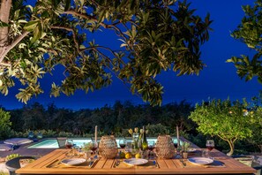 An outdoor dining table set with plates, glasses, and a bottle, surrounded by lush greenery and a pool in the background. - Giardino degli Avocadi (Giarre)
