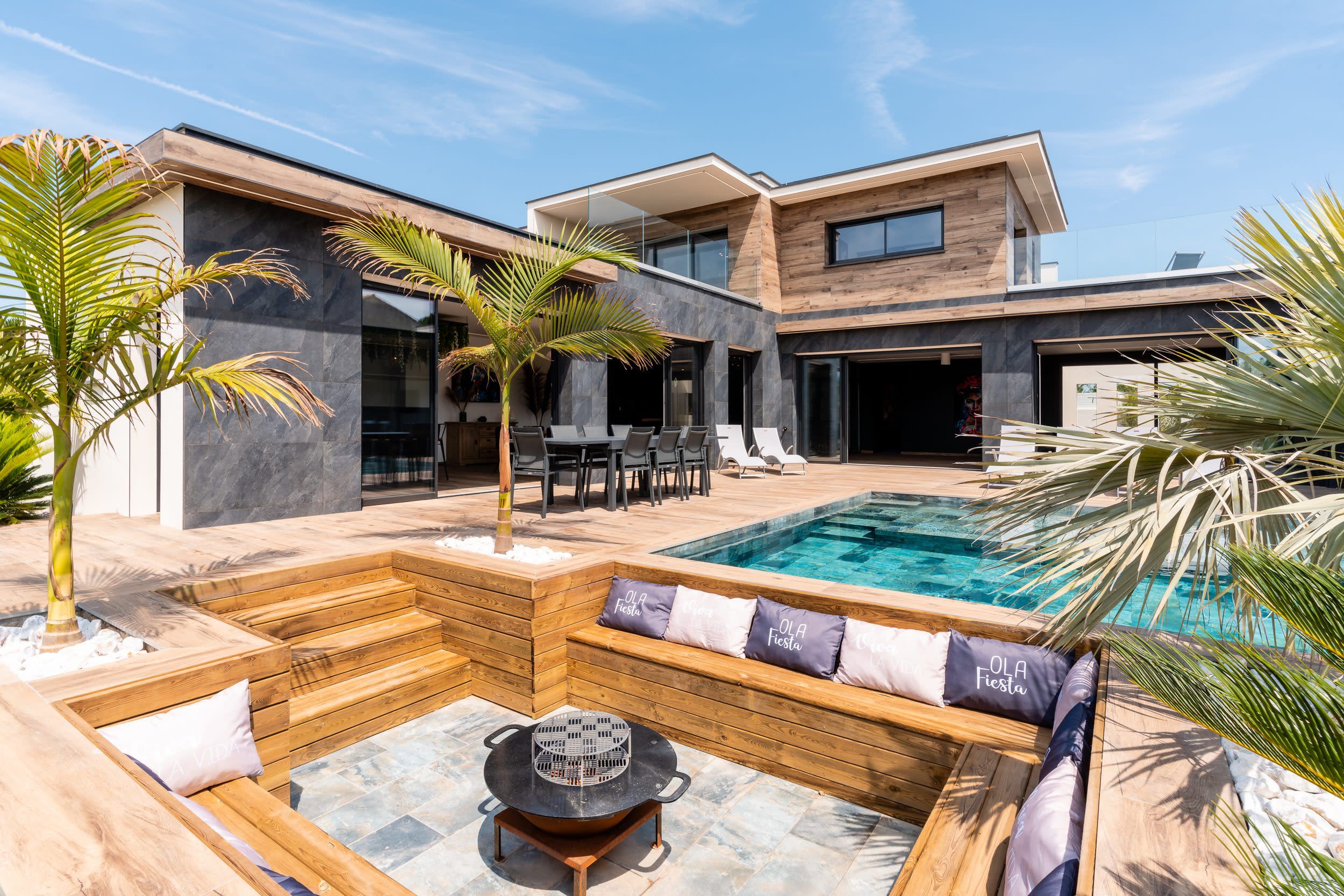 Un área de piscina moderna al aire libre con una terraza de madera, sillones de descanso y una parrilla.
