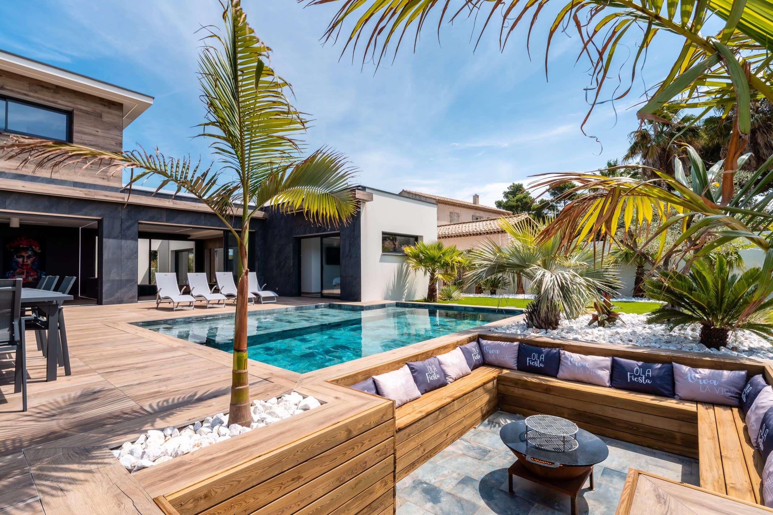 Un espace piscine extérieur moderne, doté d’une terrasse en bois, de chaises longues et d’un coin salon avec des coussins.