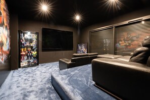 A home theater room with a large flat-screen TV, a glass display case with collectibles, a leather sofa, and a window with blinds.