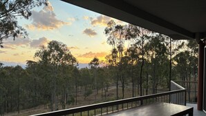 A wooden table and chairs on a balcony with a view of trees and a sunset. - Luxury new 2 bedrm with ensuites, scenic views & outdoor bath close to winery 🍇 (Mount Alford)