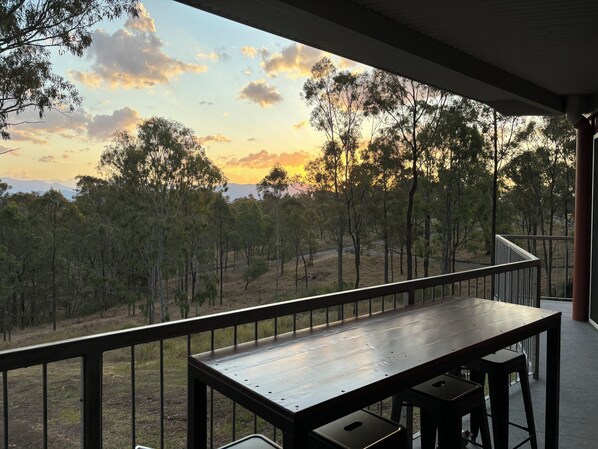 Una mesa y sillas de madera en un balcón con vista a árboles y al atardecer.
