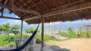 Une terrasse au bord de la plage, avec une table et des chaises en bois, un hamac et une structure dotée d’un toit de chaume.