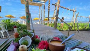 Un espace de restauration aménagé au bord de la plage, proposant une variété de plats, notamment un bol de soupe, du riz et des légumes frais, le tout sous une pergola en bois.