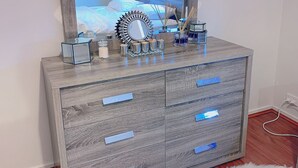 Een slaapkamer met een grote spiegel, een houten dressoir met lades, een bed met wit beddengoed, en een nachtkastje met een lamp en een vaas.