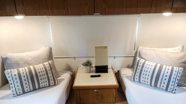 Cabine compacta e bem iluminada com duas camas, uma mesa pequena com abajur e uma planta em vaso.