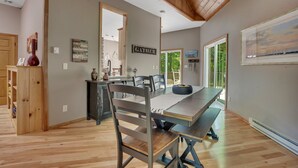 A dining area with a long table, chairs, and a bench. There is a wooden ceiling and a view of the outdoors through a glass door.
