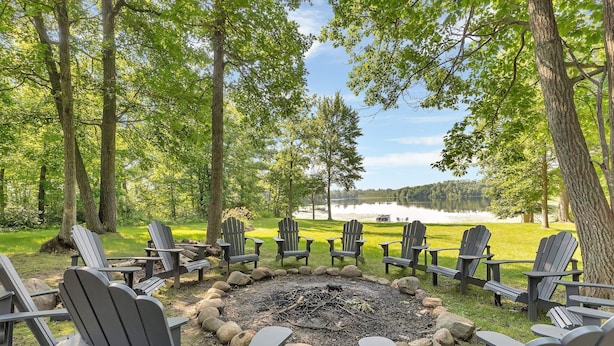 A circular seating area with Adirondack chairs around a fire pit, set in a forested area with a lake view.