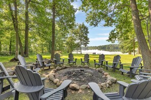 A circular seating area with Adirondack chairs around a fire pit, set in a forested area with a lake view.