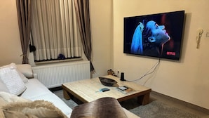 A living room with a sofa, a coffee table, and a TV mounted on the wall displaying a show. - MAISON UNIFAMILIALE (Brussels)