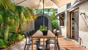 An outdoor patio with a wooden table and chairs, surrounded by lush tropical plants and a large umbrella. - Lovely 2 bedroom ground floor apartment in Noosa Heads with generous private courtyard. (Noosa Heads)