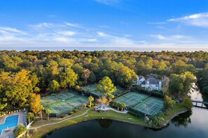 Vista aérea de un complejo de canchas de tenis rodeado de un bosque denso, una piscina y una casa grande.