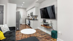Een moderne keuken met witte kasten, een roestvrijstalen koelkast en een magnetron. Er is een flatscreen televisie aan de muur bevestigd, een glazen plank met boeken, en een ronde witte tafel.