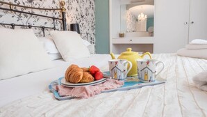 Un lit bien fait, sur lequel on a posé un plateau avec des croissants et des fraises, deux tasses décorées avec un motif de maison, et une théière jaune sur un couvre-lit blanc.