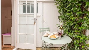 Un petit espace extérieur aménagé avec une table, des chaises, un livre, un verre de vin rouge et une bouteille de vin.