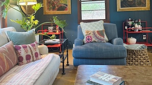 Una sala de estar con sofá, sillón, mesa de centro y estante metálico rojo con libros y objetos decorativos.