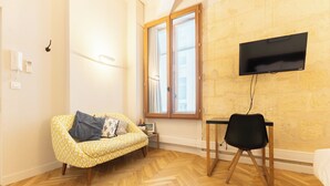 Una habitación con sofá, un escritorio y una silla, televisor, ventana con persianas y piso de madera en diseño espiga.