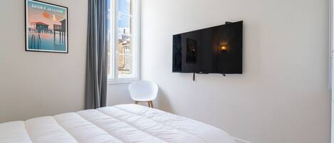 Un dormitorio con cama, televisor montado en la pared, ventana con cortinas e imagen enmarcadas en la pared.