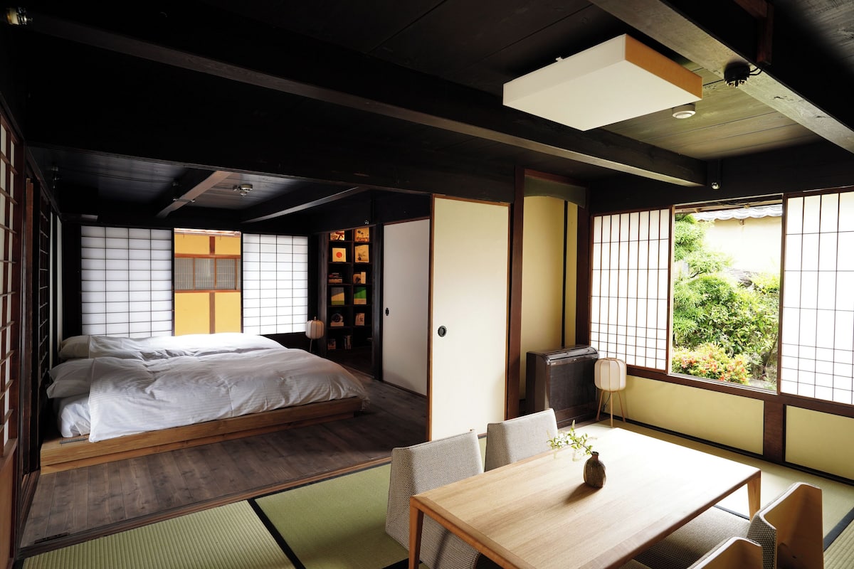 Ein traditioneller japanischer Raum mit Tatami-Boden, einem niedrigen Holzbett, einem Tatami-Tisch mit einer Vase und Blick ins Grüne durch Shoji-Schirme.