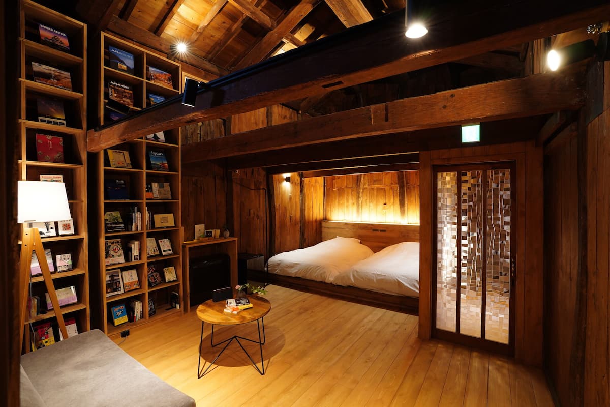 Ein gemütliches Zimmer mit Holzbalkendecke, einer Bücherwand, einem Bett und einem kleinen runden Tisch.