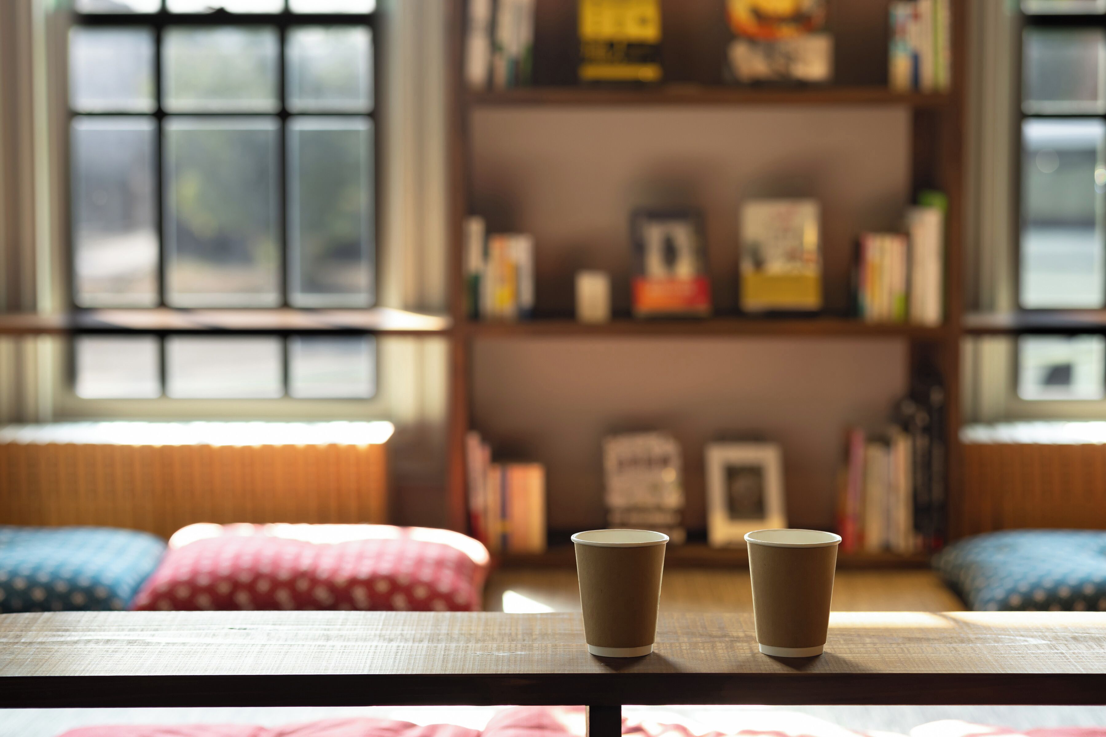 A cozy reading nook with a wooden table, two paper cups, and a bookshelf filled with books and decorative items.
