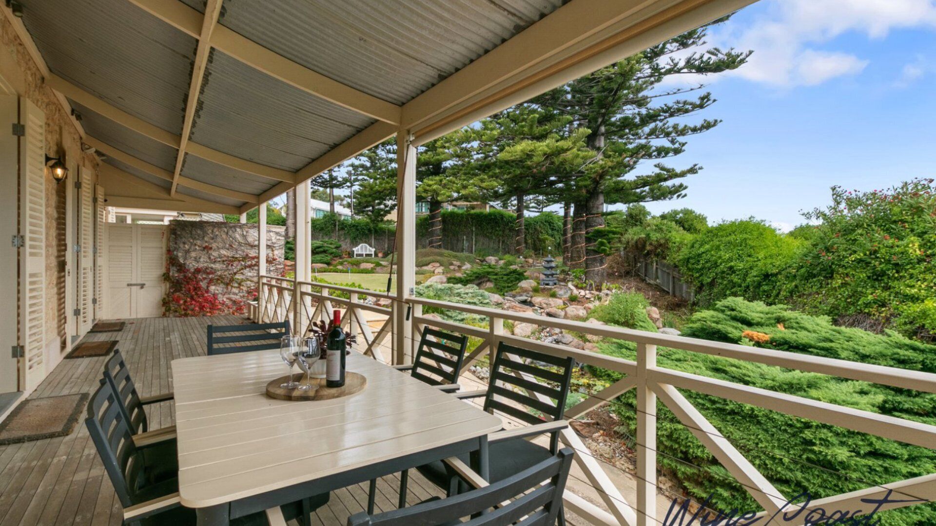Un espace extérieur abrité, aménagé pour les repas, avec une table et des chaises, donnant sur un jardin paysager agrémenté d’arbres et d’arbustes.