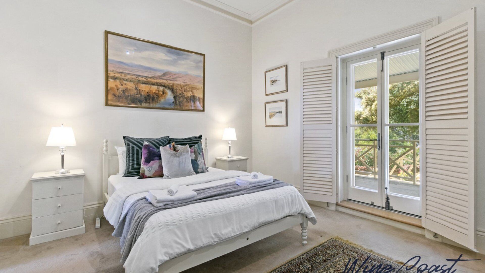 A bedroom with a bed, two bedside tables with lamps, a large window with shutters, and a painting above the bed.
