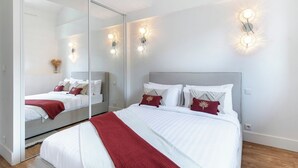 A modern bedroom with a large mirror, a bed with white linens and red accents, and wall-mounted lighting. - Penthouse Massena - 2 bedroom apartment (Nice)