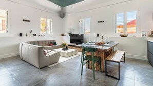 Een ruime woonkamer met een bank, salontafel en eettafel. Een televisie is aan de muur bevestigd, en de kamer heeft grote ramen.