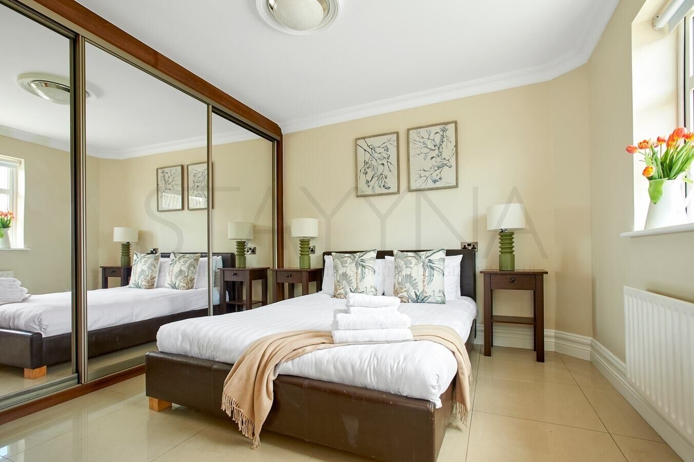 A hotel bedroom with a large mirror, two beds, a bedside table with a lamp, and artwork on the wall.