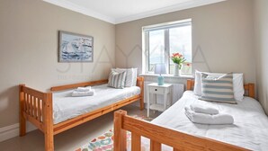 A bedroom with two wooden beds, a window with a view, a small table with a lamp, and a painting on the wall. - Beachfront Break - 2 bedroom home (County Kerry)