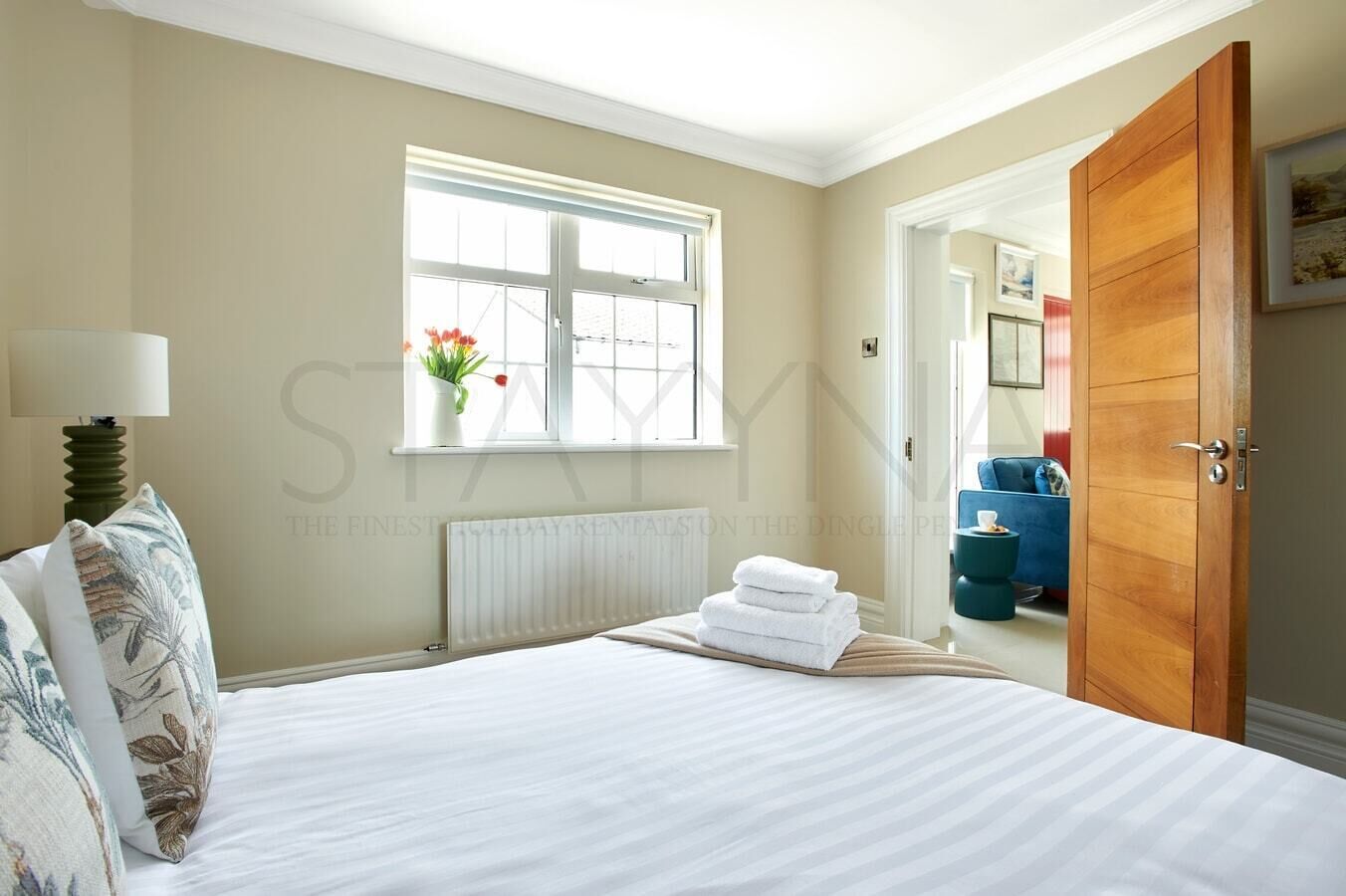 A hotel room with a large bed, a radiator, a window with flowers, a blue chair, and a wooden door.