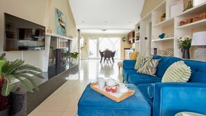 A spacious living room with a blue sofa, a coffee table with a tray of pastries, a TV, and a kitchen in the background. - Beachfront Break - 2 bedroom home (County Kerry)