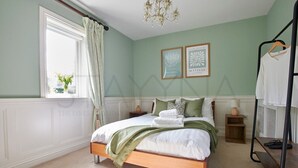 Een slaapkamer met een bed, een raam met gordijnen, een kroonluchter en twee ingekaderde afbeeldingen aan de muur.