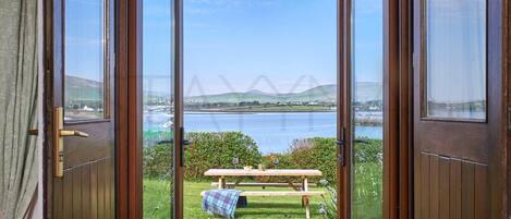 A view through a wooden door with a glass panel, overlooking a grassy area with a picnic table and a bench, and a body of water with a bridge in the distance.