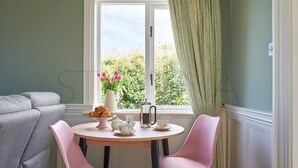 Een eethoek met een ronde tafel, twee roze stoelen, een grijze bank en een raam met groene gordijnen.
