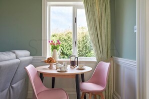 A dining area with a round table, two pink chairs, a grey sofa, and a window with green curtains. - Waterfront Annex one bedroom annex (Dingle)
