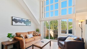 A loft-style living room with a high ceiling, a large window, a brown leather sofa, a wooden coffee table, and a floor lamp.