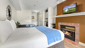 A bedroom with a bed, a television on a shelf above a fireplace, and a kitchen in the background. - Comfort Studio - 210B (Kelowna)