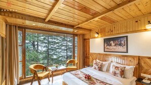 A wooden cabin bedroom with a large window, a bed with floral bedding, a chair, and a table with a book and flowers. - Zostel Kotgarh (Narkanda) (Kumharsain)