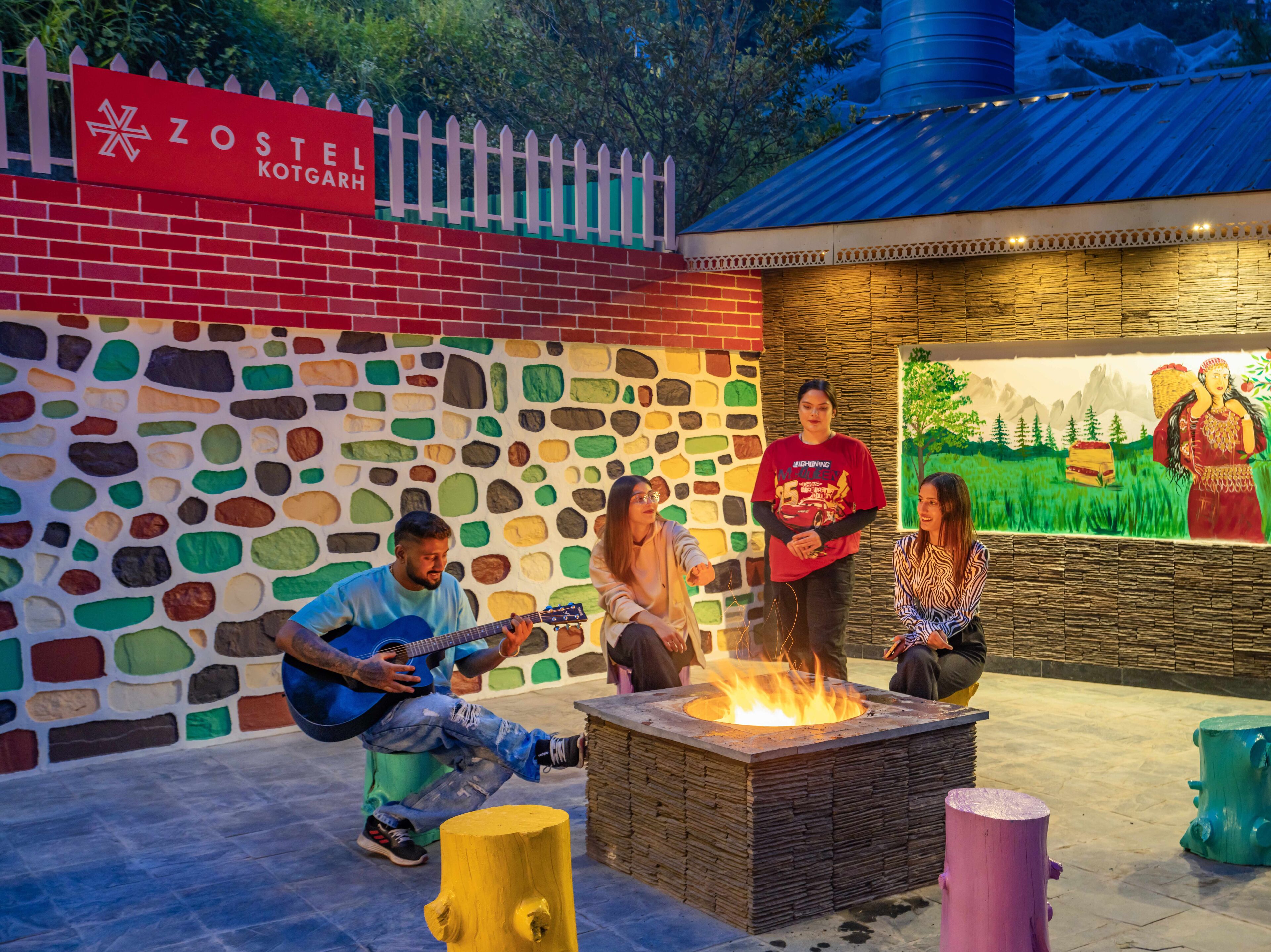 Un grupo de personas sentadas alrededor de una fogata, con un muro de piedra y un mural al fondo.