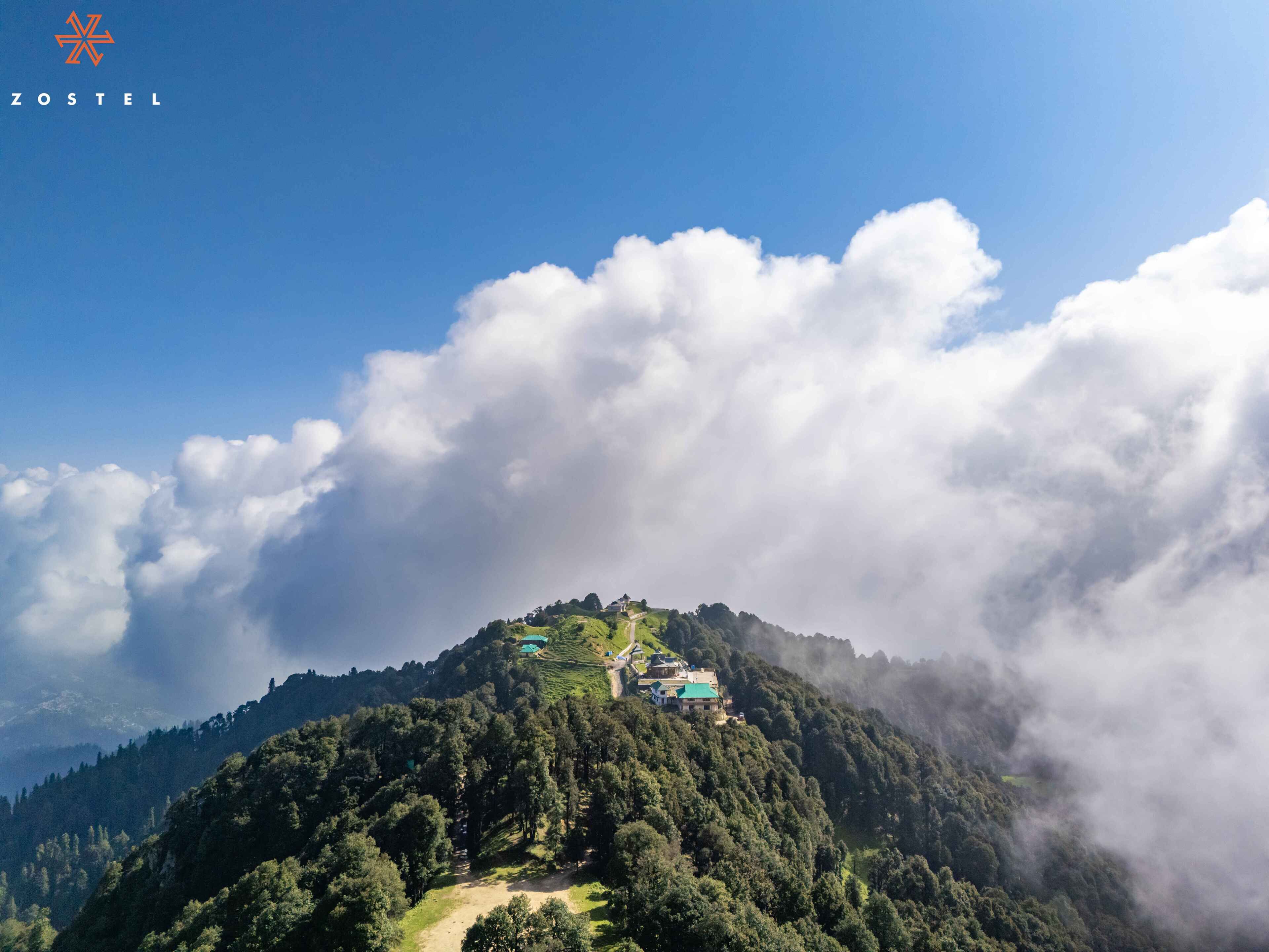 A mountainous landscape with a small cluster of buildings on a hilltop, surrounded by dense forest and clouds.