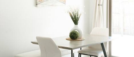 A dining area with a round table, white chairs, a vase with plants, and a painting on the wall.