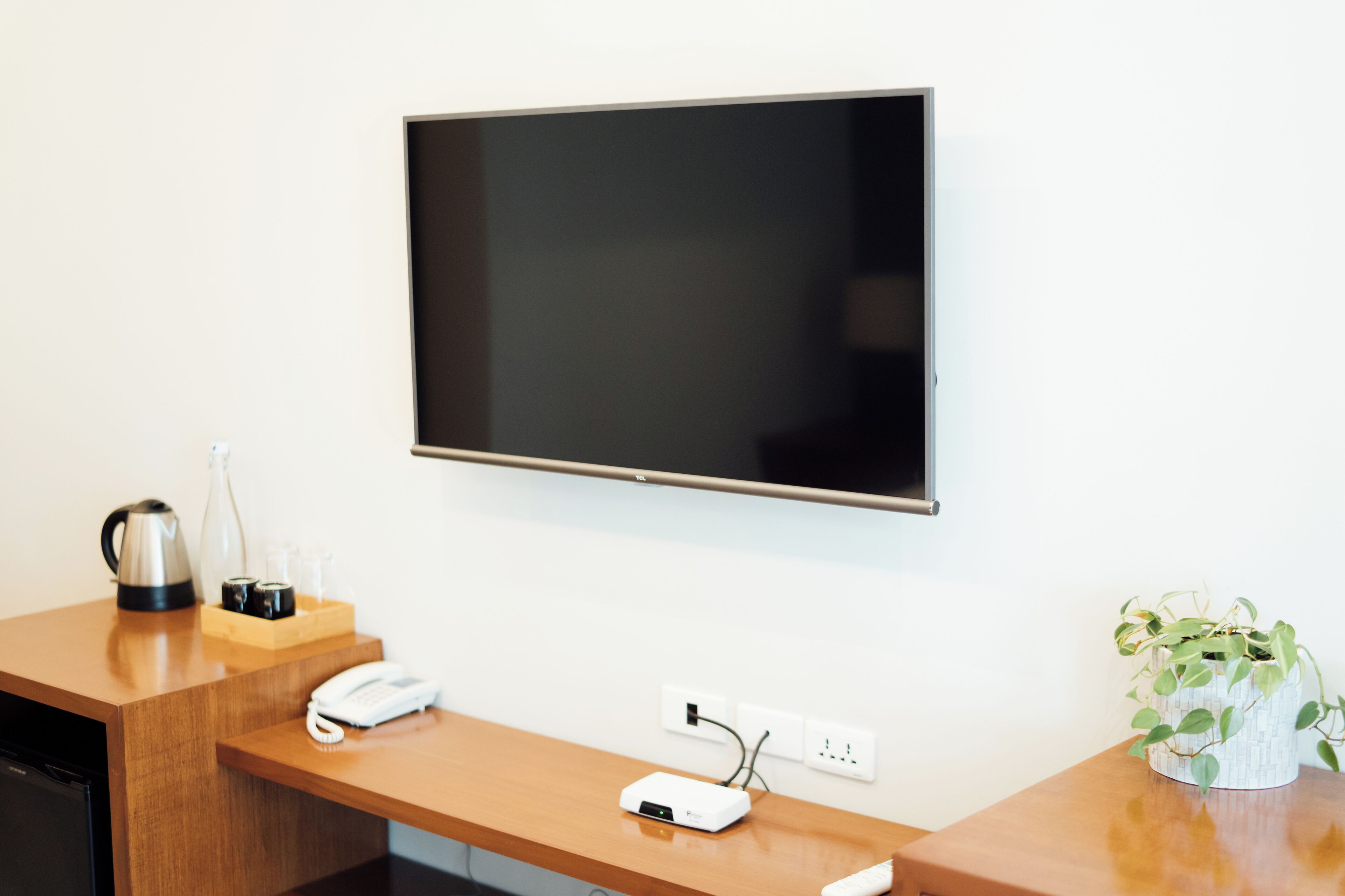 Habitación de hotel con televisor de pantalla plana montado en la pared, un escritorio de madera con teléfono, cafetera y regleta de enchufes.